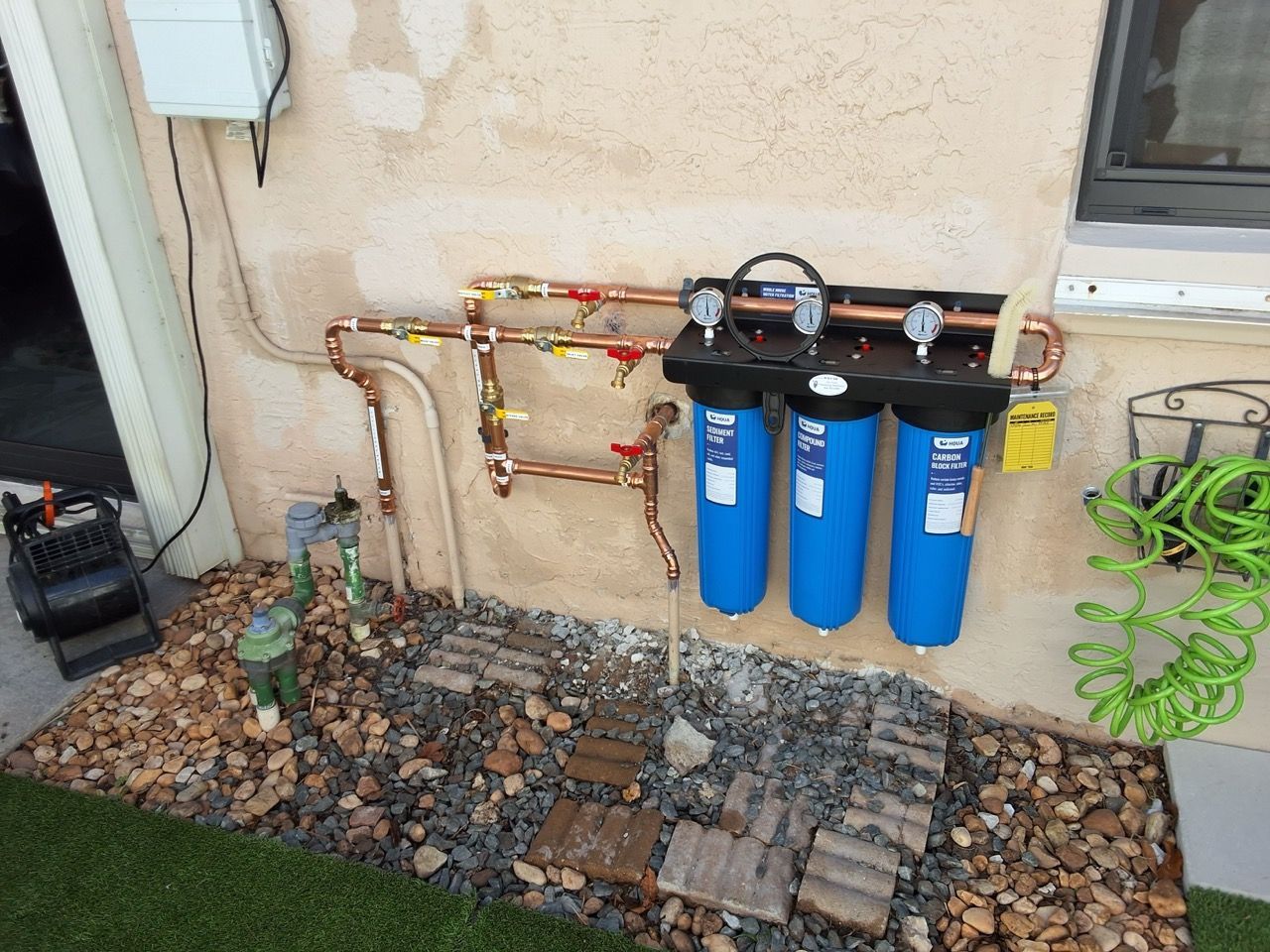 Water filtration system with blue canisters and copper pipes beside a stucco wall in a rock garden.