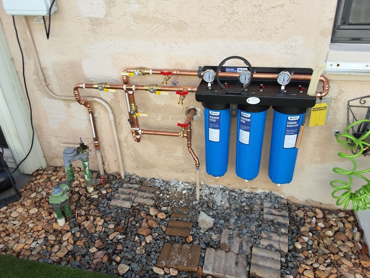 Three blue water filter canisters mounted on an exterior wall with copper pipes and gravel below