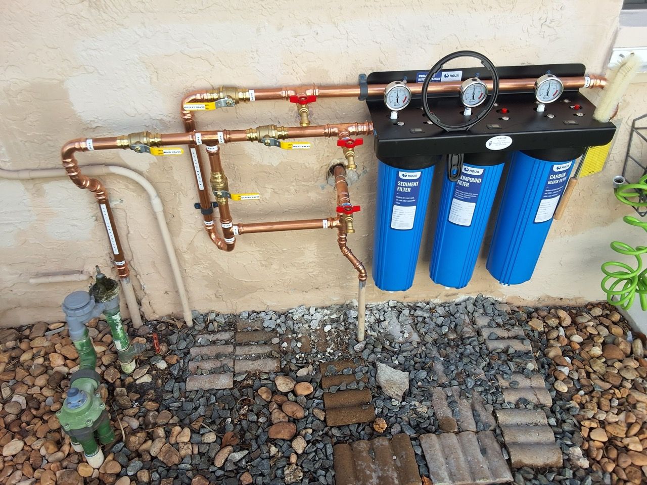 Outdoor water filtration system with copper piping and three blue filters mounted on a wall.