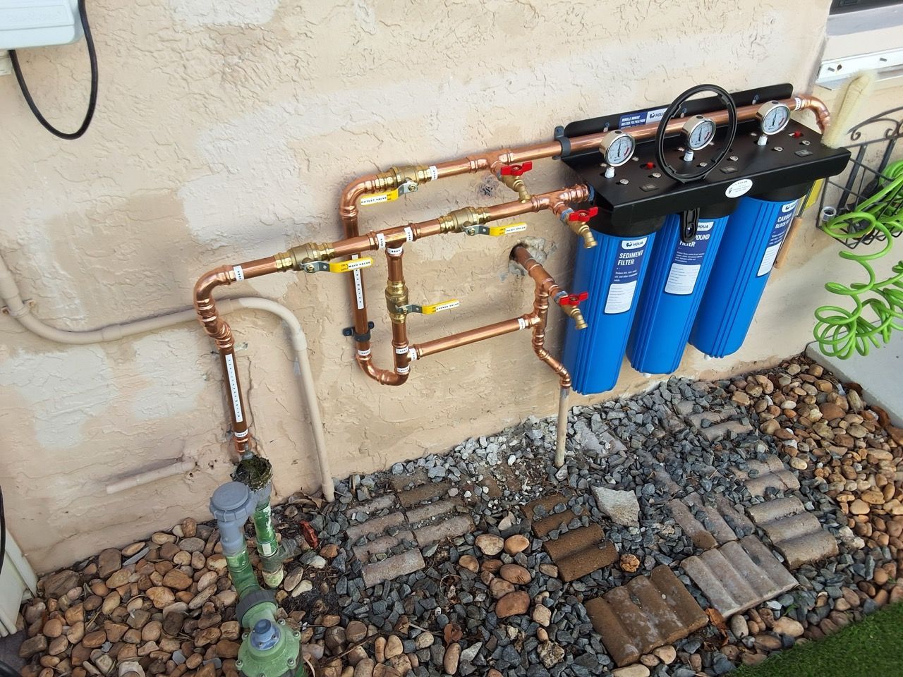 Outdoor water filtration setup with copper pipes and three blue canister filters against a stucco wall.
