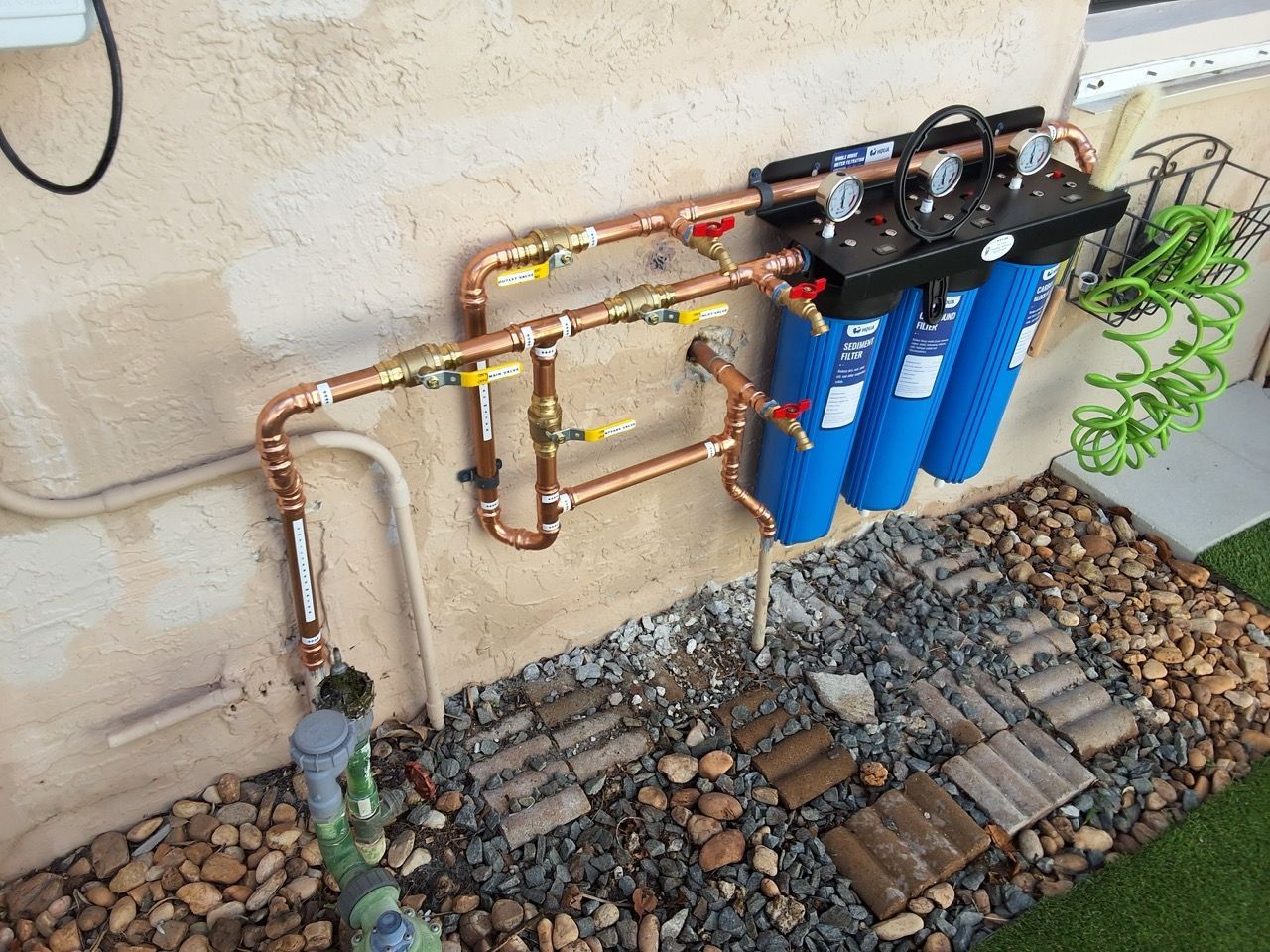 Copper water filtration system with blue cylinders mounted on an outdoor wall over gravel.