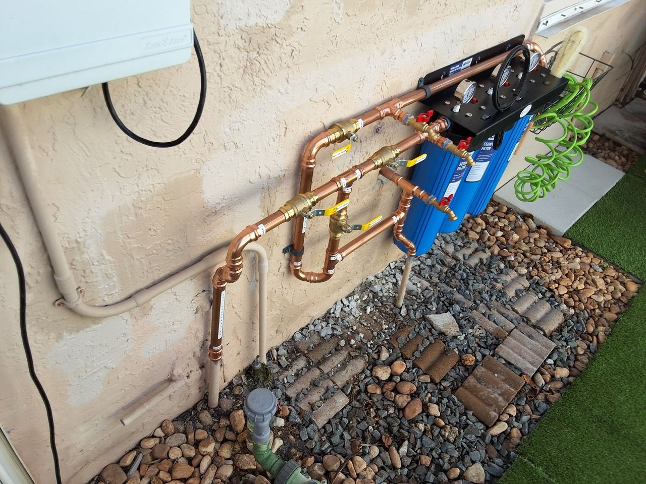 Outdoor water pipes and valves mounted on a stucco wall beside gravel landscaping and a blue tank