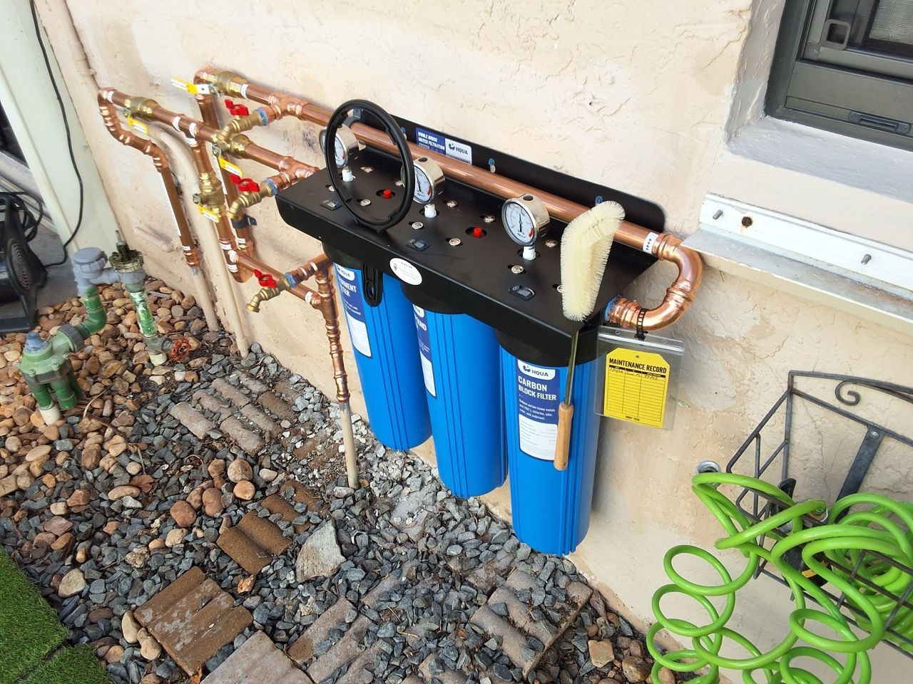 Water filter system with three blue canisters mounted on an exterior wall beside copper pipes and gravel.