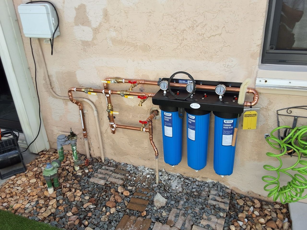 Three blue water filter canisters mounted on an exterior wall with copper piping and valves