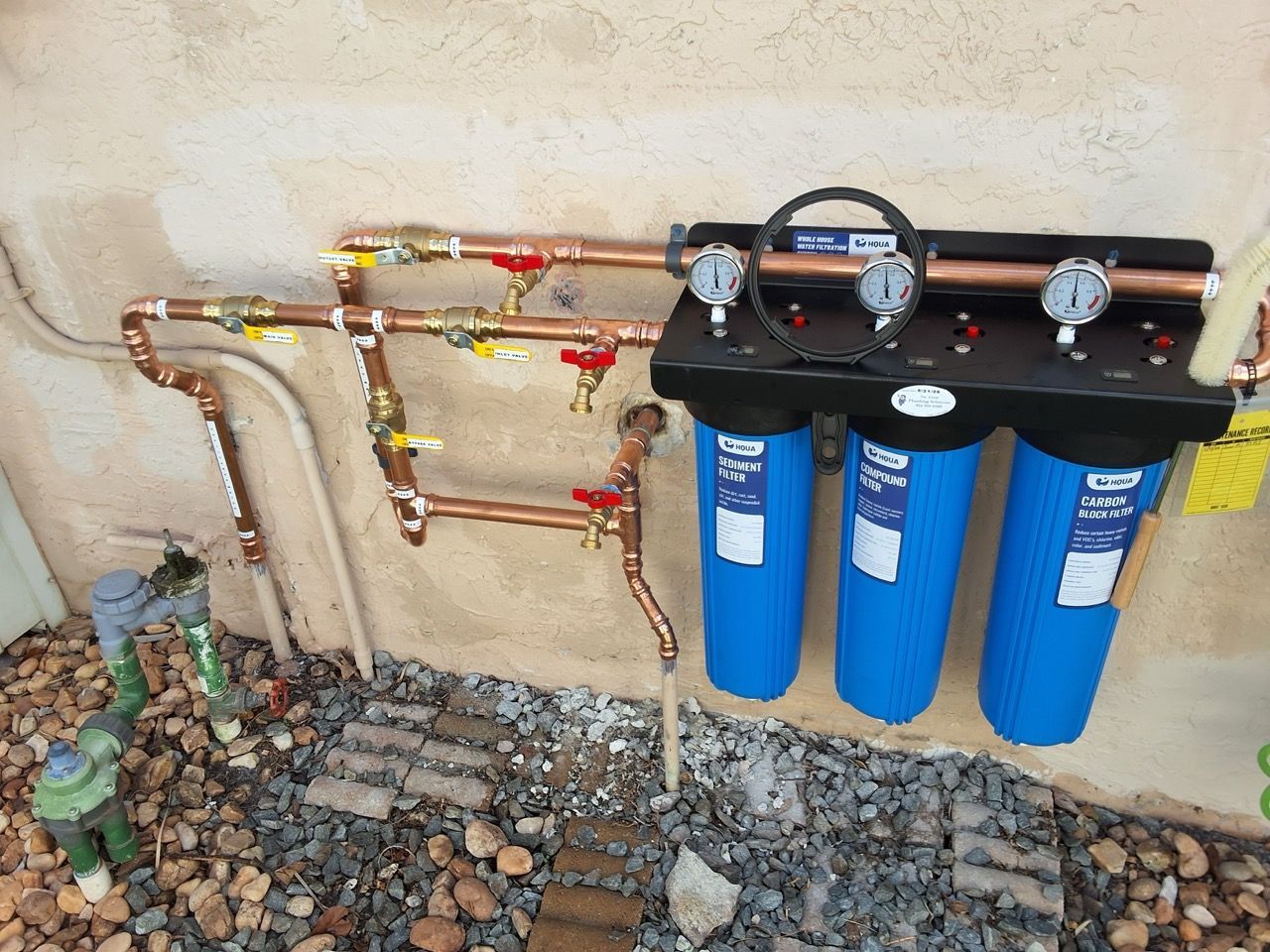 Blue water filter system with copper pipes mounted on an exterior wall, above gravel.