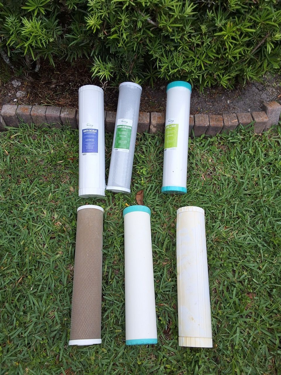 Six rolled mats or tubes in white, tan, and green, laid out on grass near a hedge.