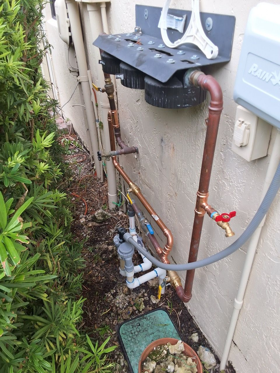 Outdoor utility pipes and valves beside a house wall, with a shrub on the left.
