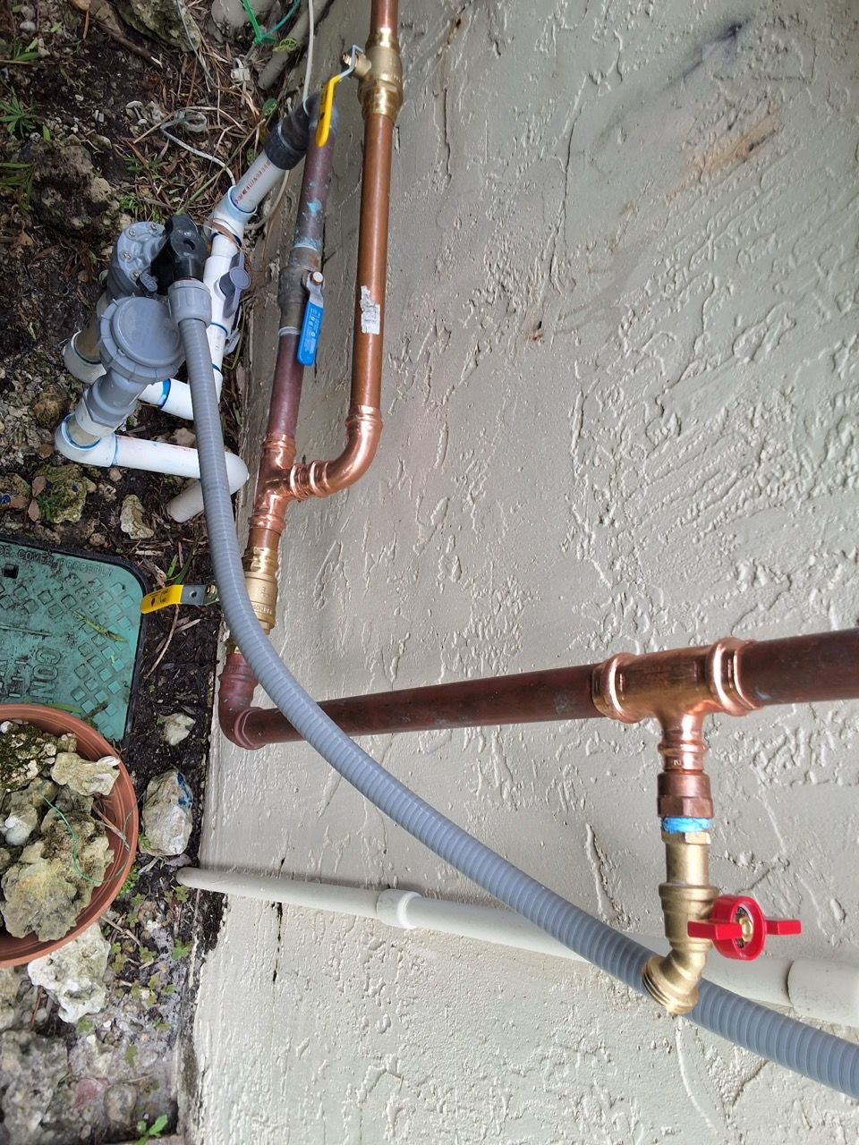 Outdoor copper and gray PVC pipes with a red valve beside a wall and ground debris.