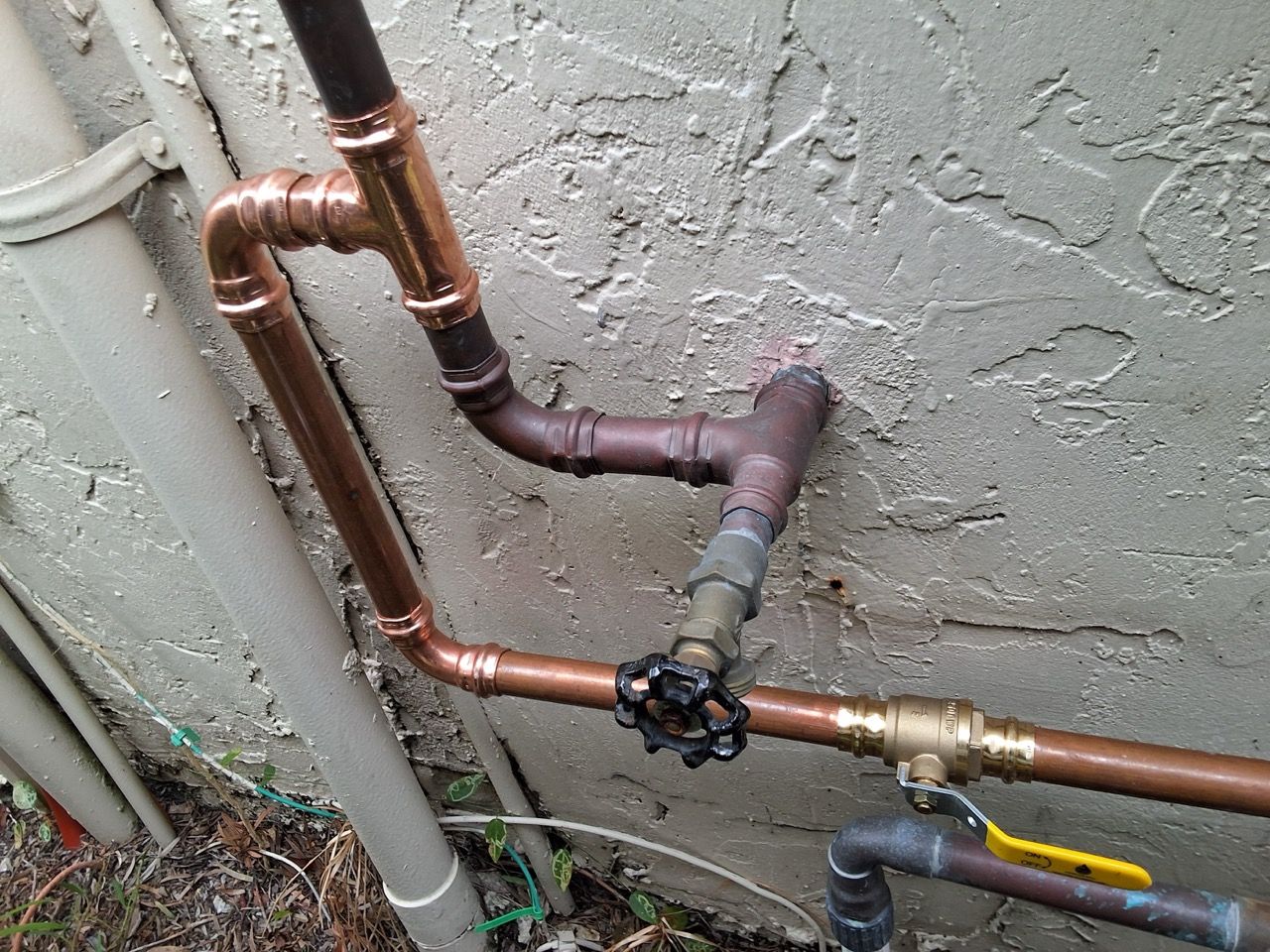 Copper and PVC plumbing pipes with a black valve outdoors against a stucco wall.