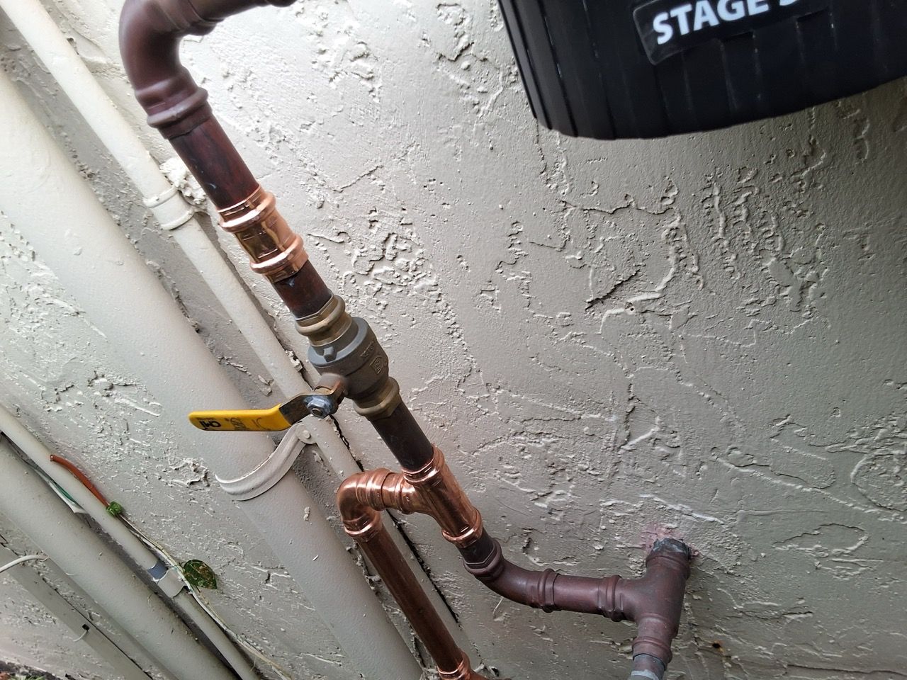 Copper and PVC pipes with a yellow-handled valve against a textured concrete wall.