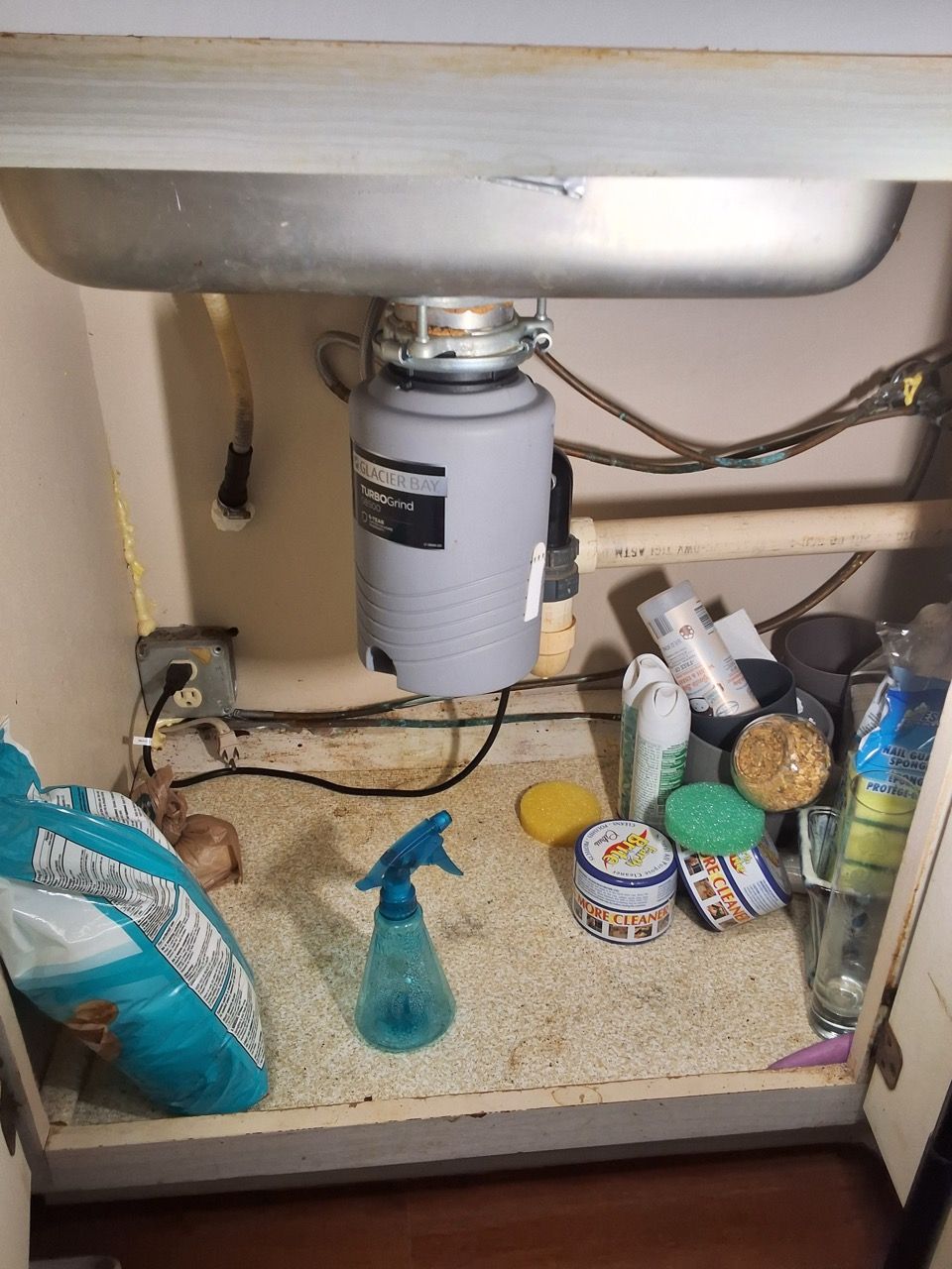 Under-sink cabinet with plumbing, cleaning supplies, and a cluttered shelf.