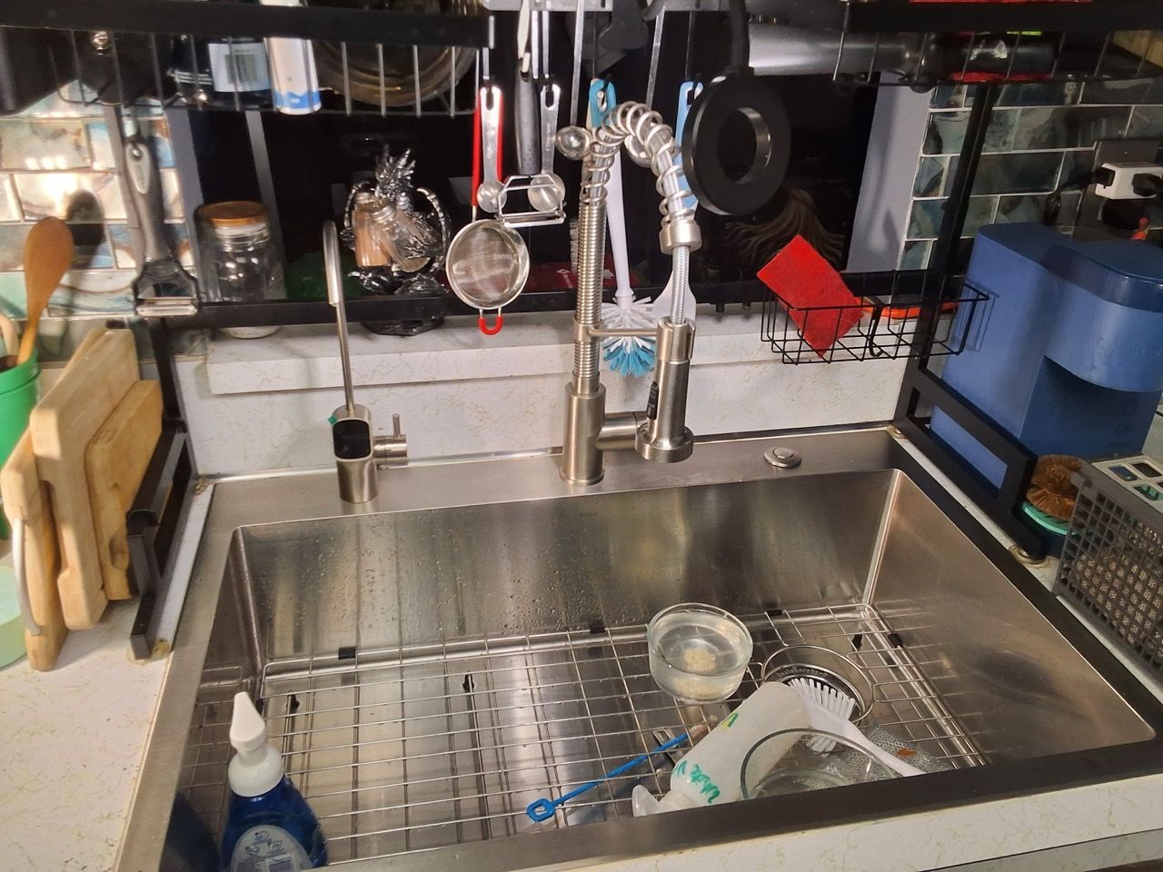 Stainless steel kitchen sink under a cluttered counter with soap bottle, dishes, and hanging utensils