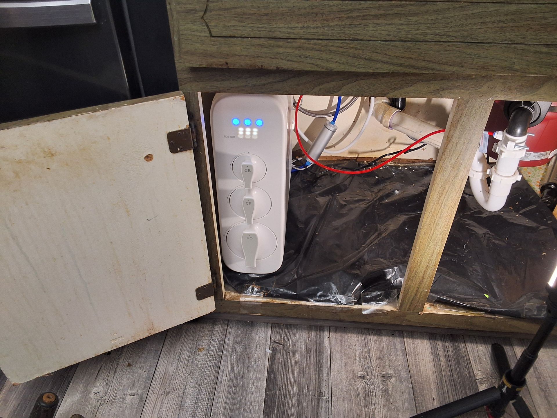 White portable appliance under a cabinet with blue lights, beside exposed wiring and pipes on a carpeted floor