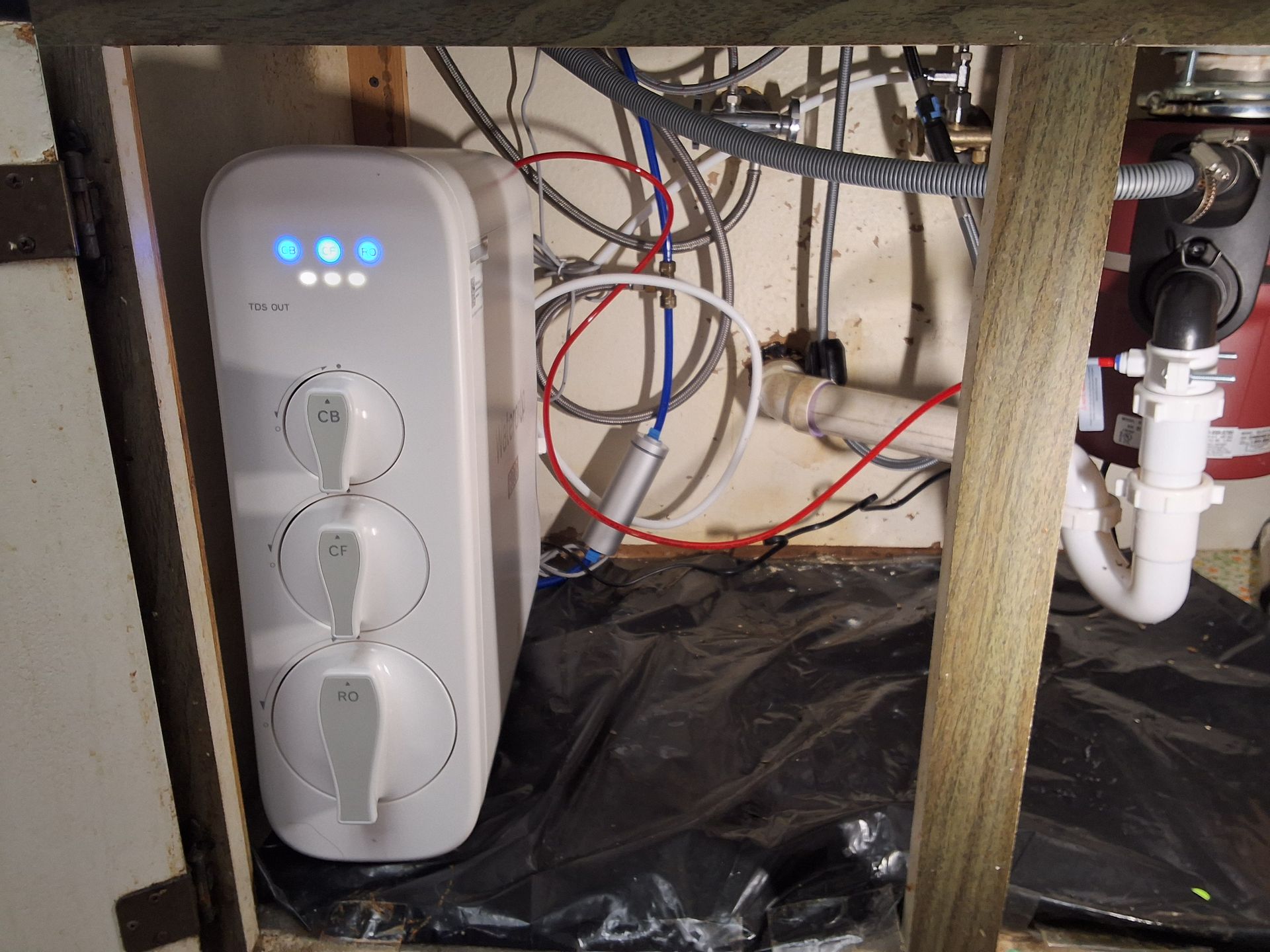 White three-outlet power controller with blue lights mounted under a sink amid exposed pipes and wires