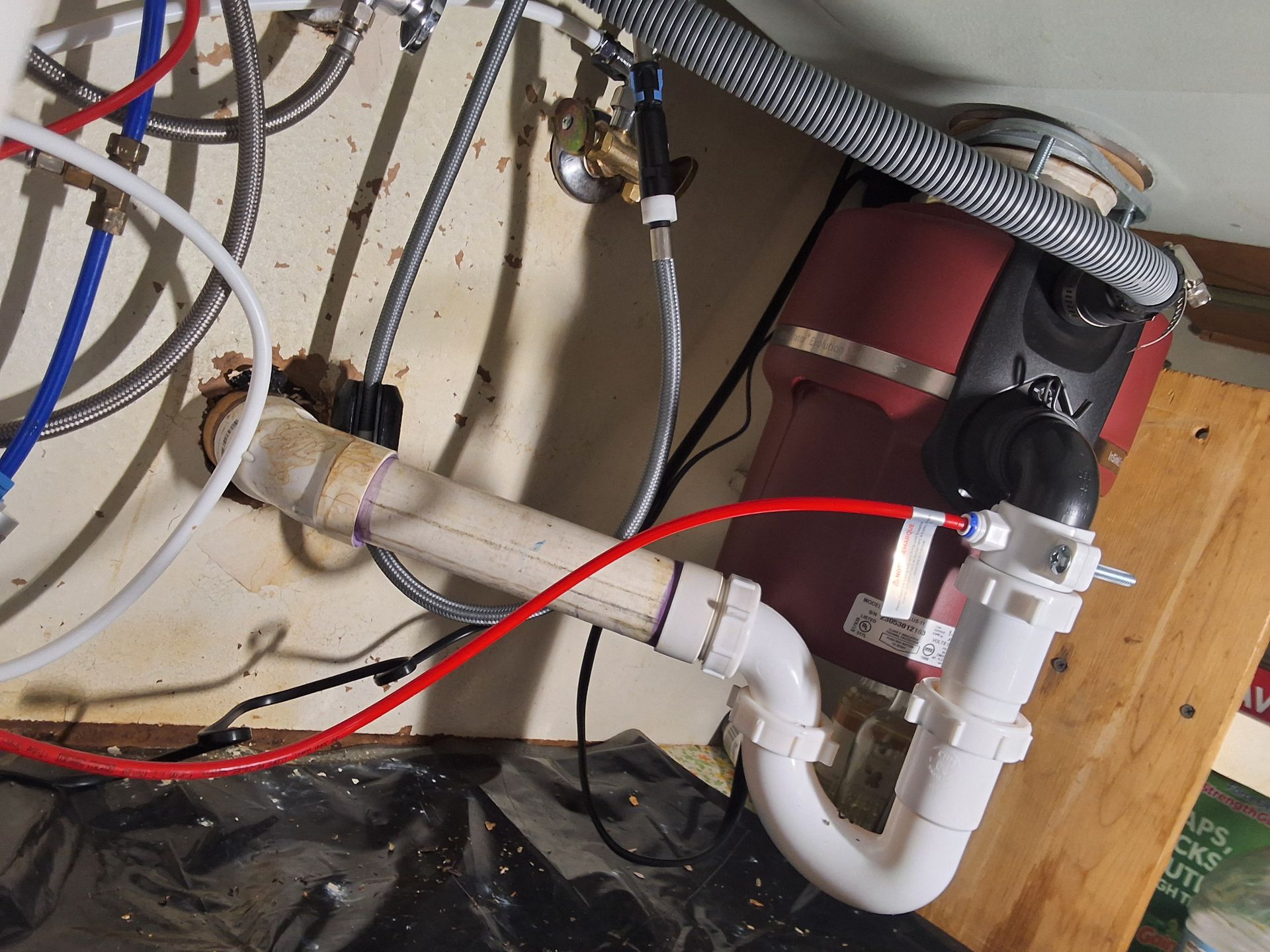 Under-sink plumbing with white PVC pipes, red and black hoses, and a red tank on a wooden wall panel