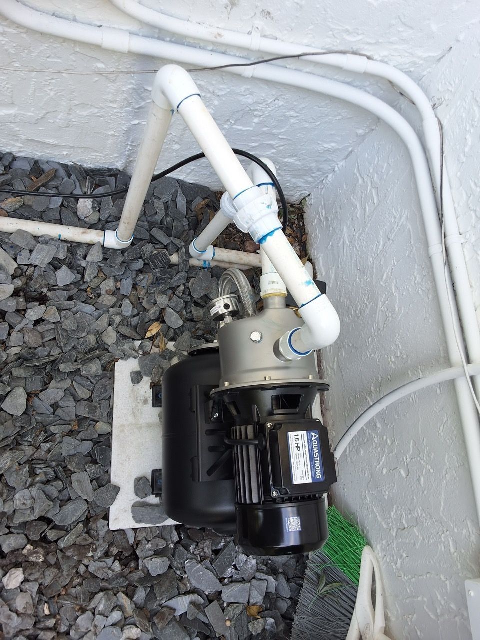 Black water pump with white PVC pipes mounted on gravel beside a wall