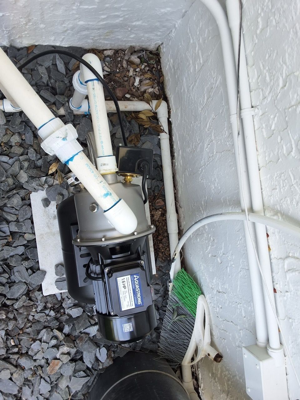 Black water pump with white PVC pipes in a gravel utility pit beside a wall.
