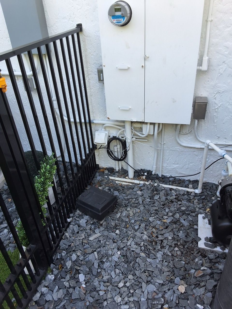 Utility meter box and electrical panels mounted on an exterior wall above gravel beside a black metal fence.