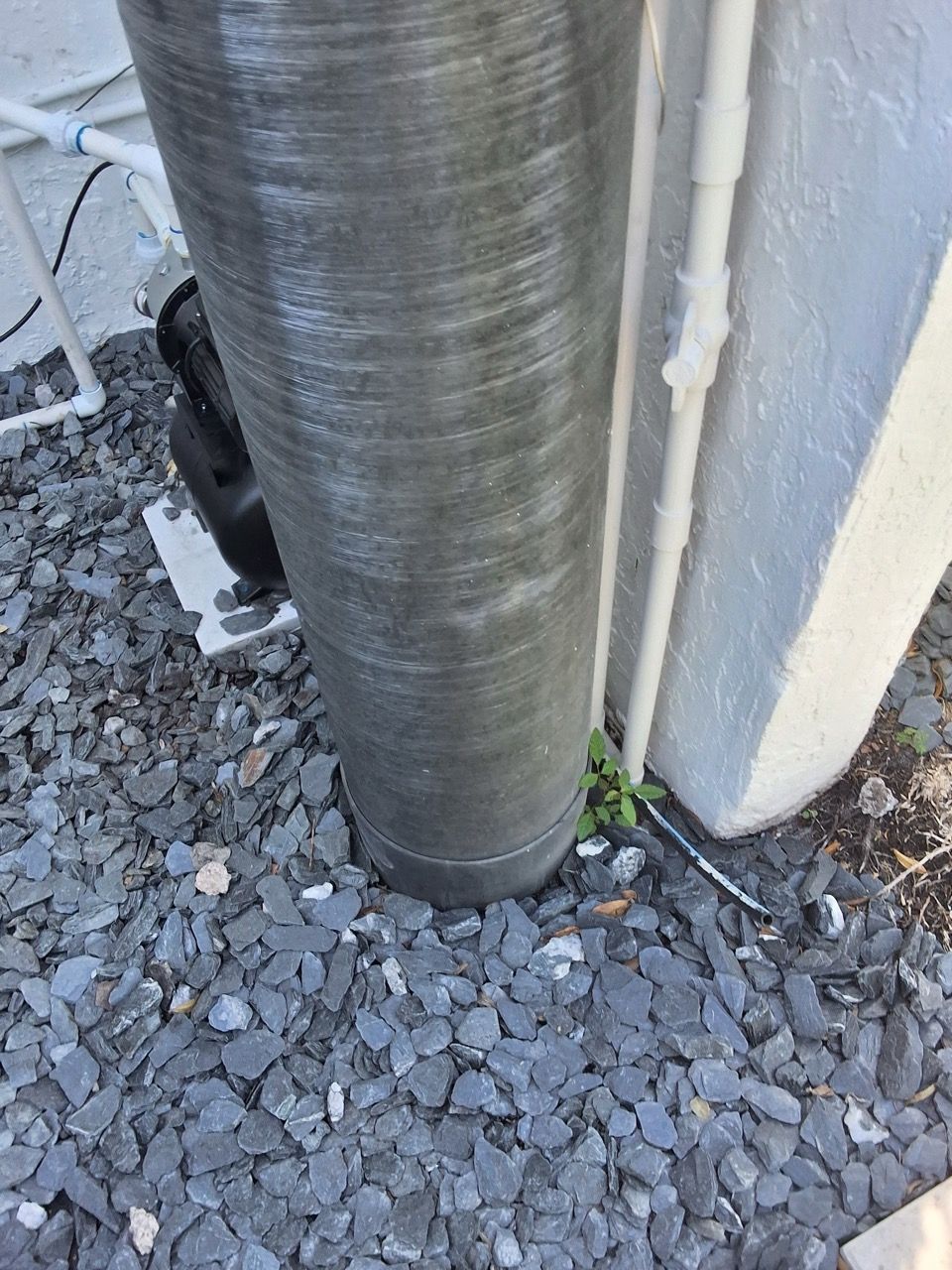 Metal pipe beside white wall on gravel ground, with small debris near the base.