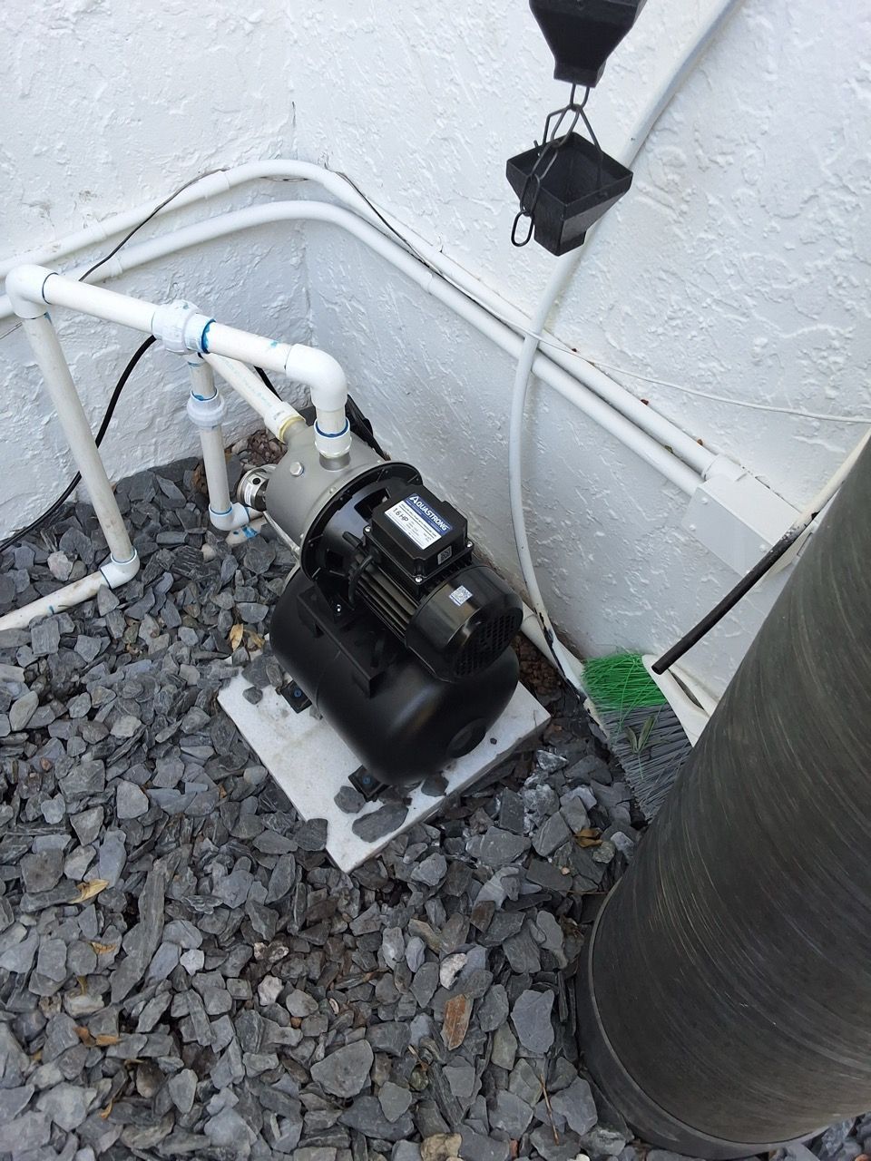 Outdoor water pump and white pipes beside a wall on gravel, next to a large gray tank.