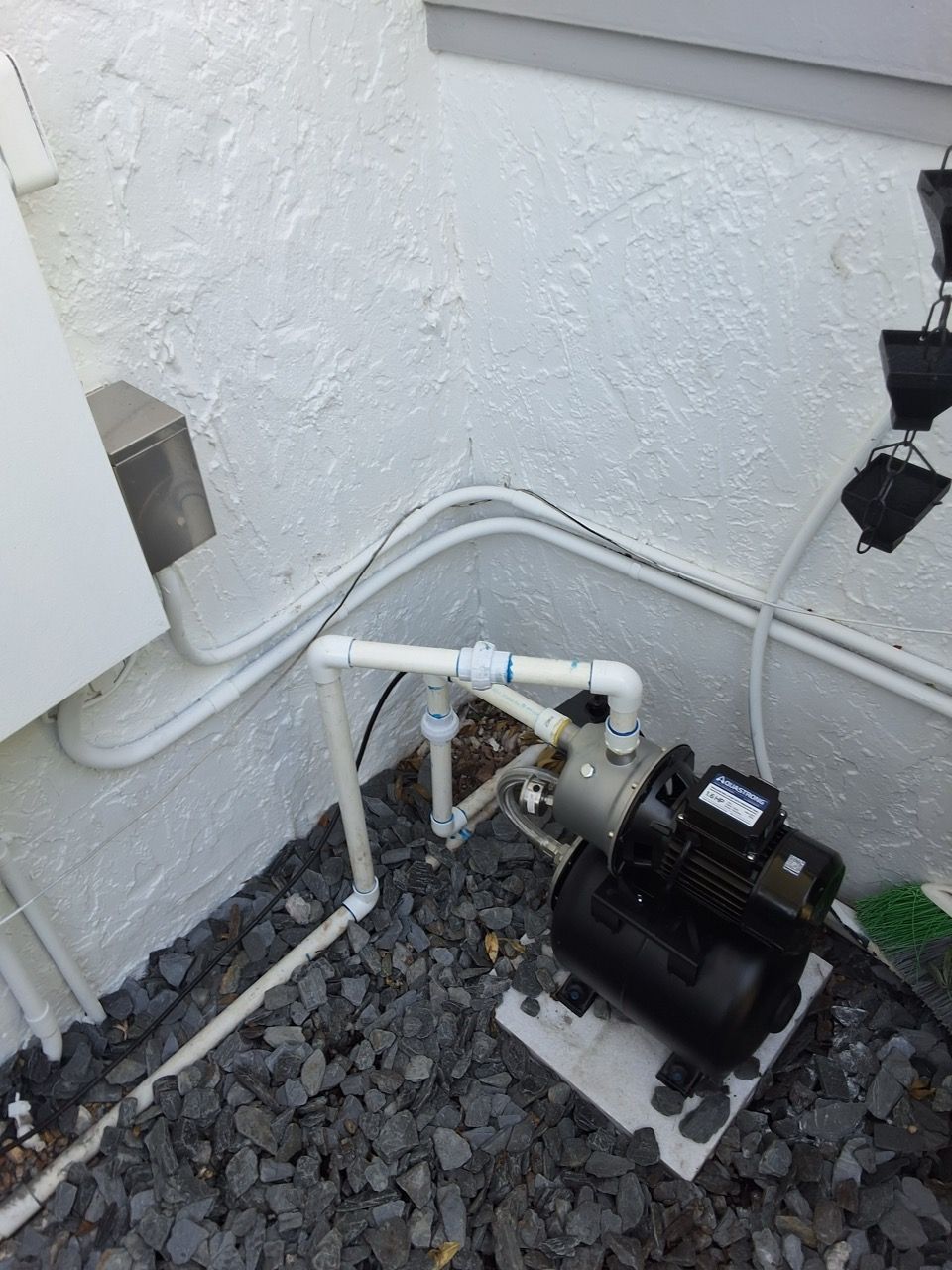 Outdoor pool pump and plumbing equipment beside a white wall on gravel.