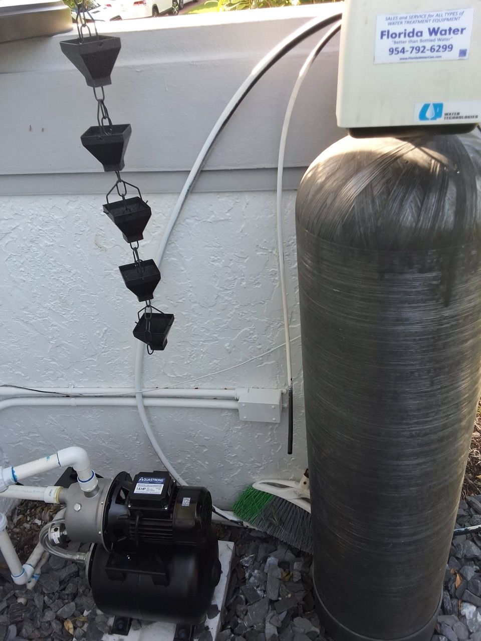 Outdoor water softener and pump setup beside a house wall, with black sediment filter and hoses.