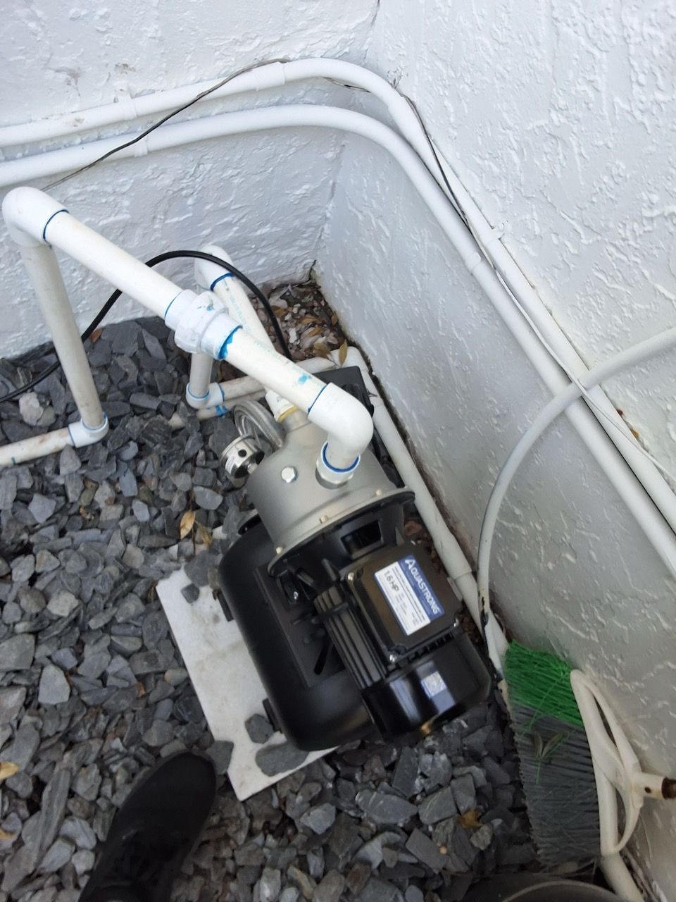 Small black water pump with white PVC pipes in a gravel-lined corner beside a wall.
