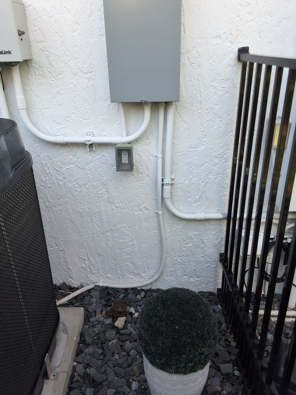Narrow outdoor utility area with electrical box, white pipes, gravel, and a black metal gate.