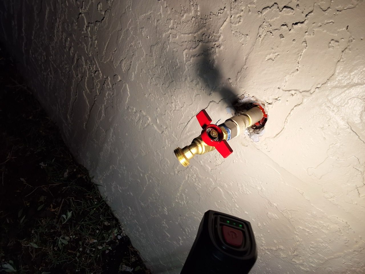 Red-handled outdoor water spigot mounted on a stucco wall at night, lit by a flashlight.