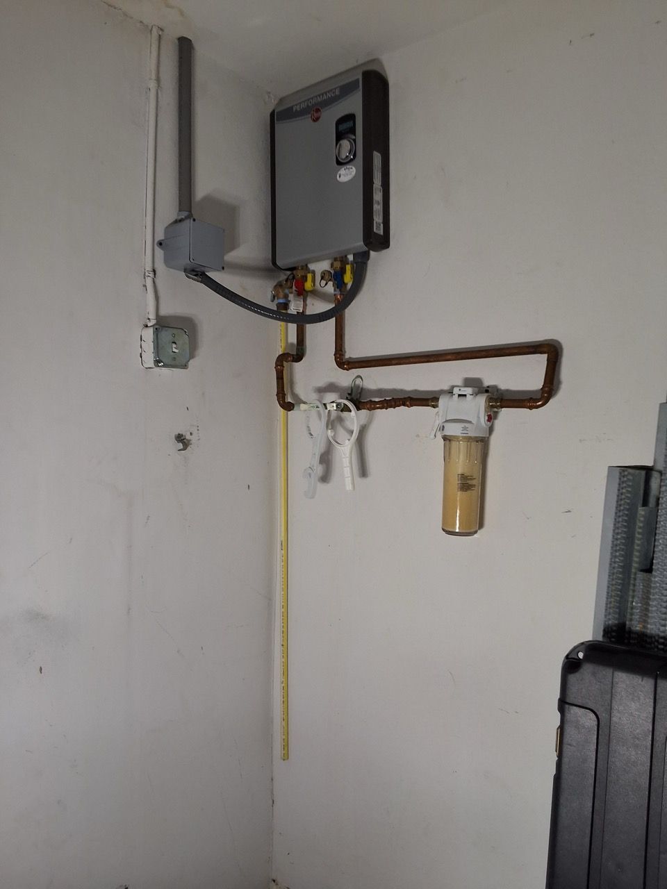 Wall-mounted water heater with pipes, valves, and a small filter in a utility room corner.