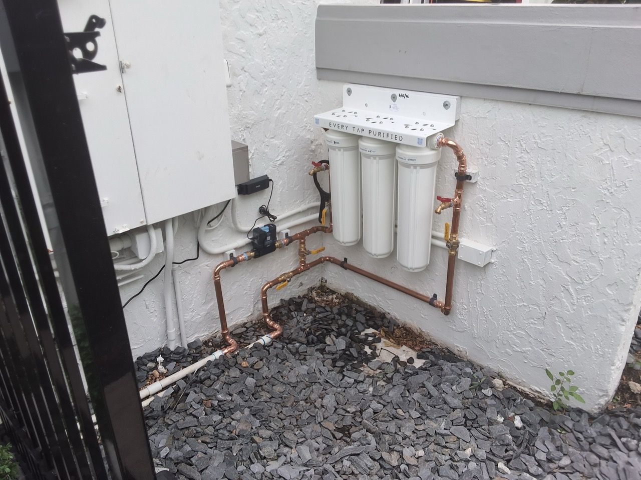 Outdoor utility meters and copper piping mounted on a white wall beside a gravel ground