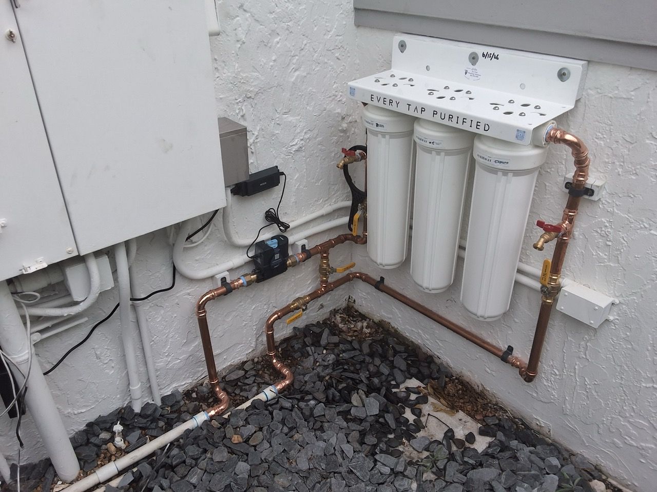 Outdoor water filtration system with three white filter cartridges, copper pipes, and a wall-mounted unit in a utility area
