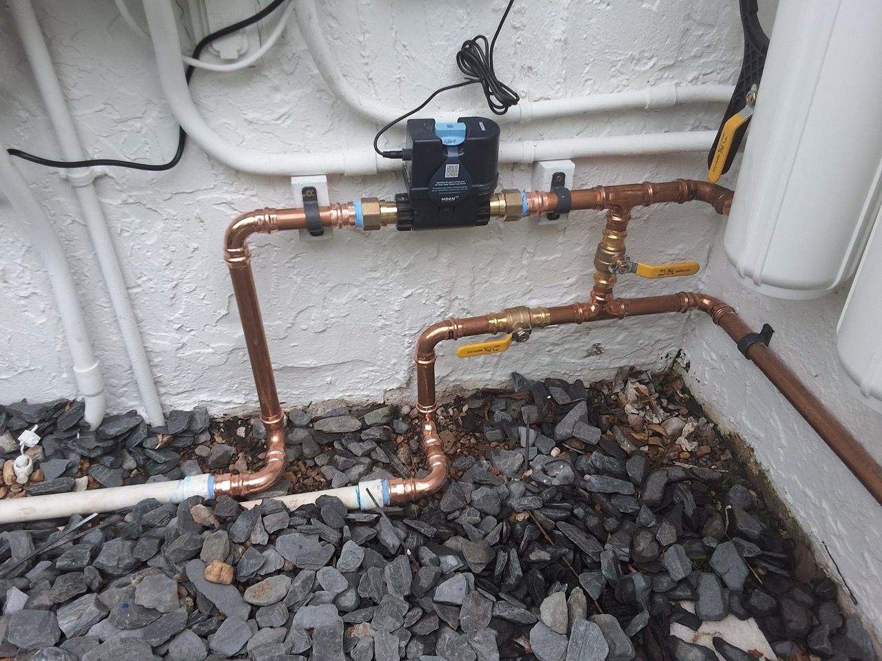 Copper pipes and yellow shutoff valves on a white wall beside a black device and gravel ground