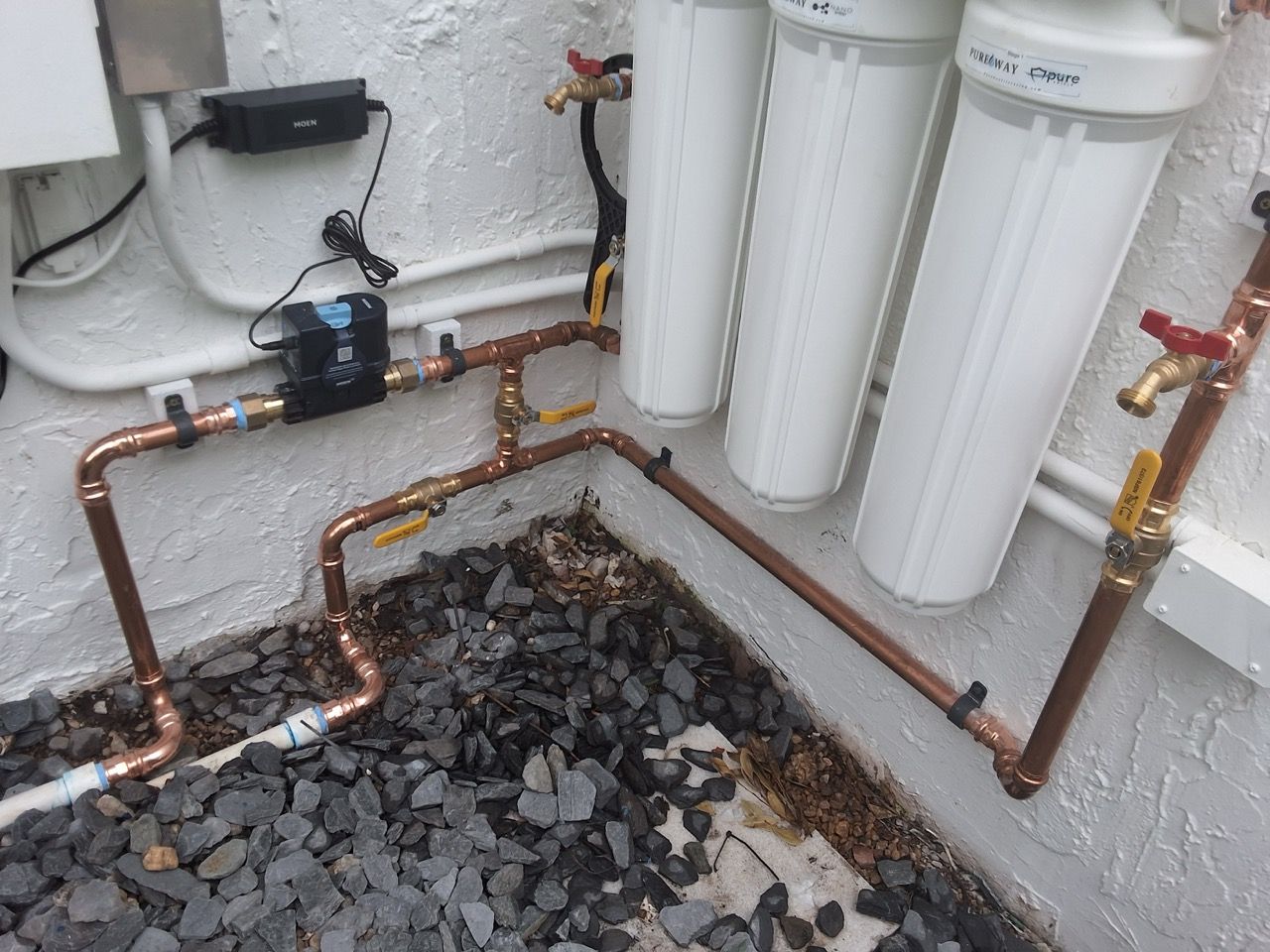 Indoor utility area with white pipes, copper plumbing, filter canisters, and a gravel floor trench