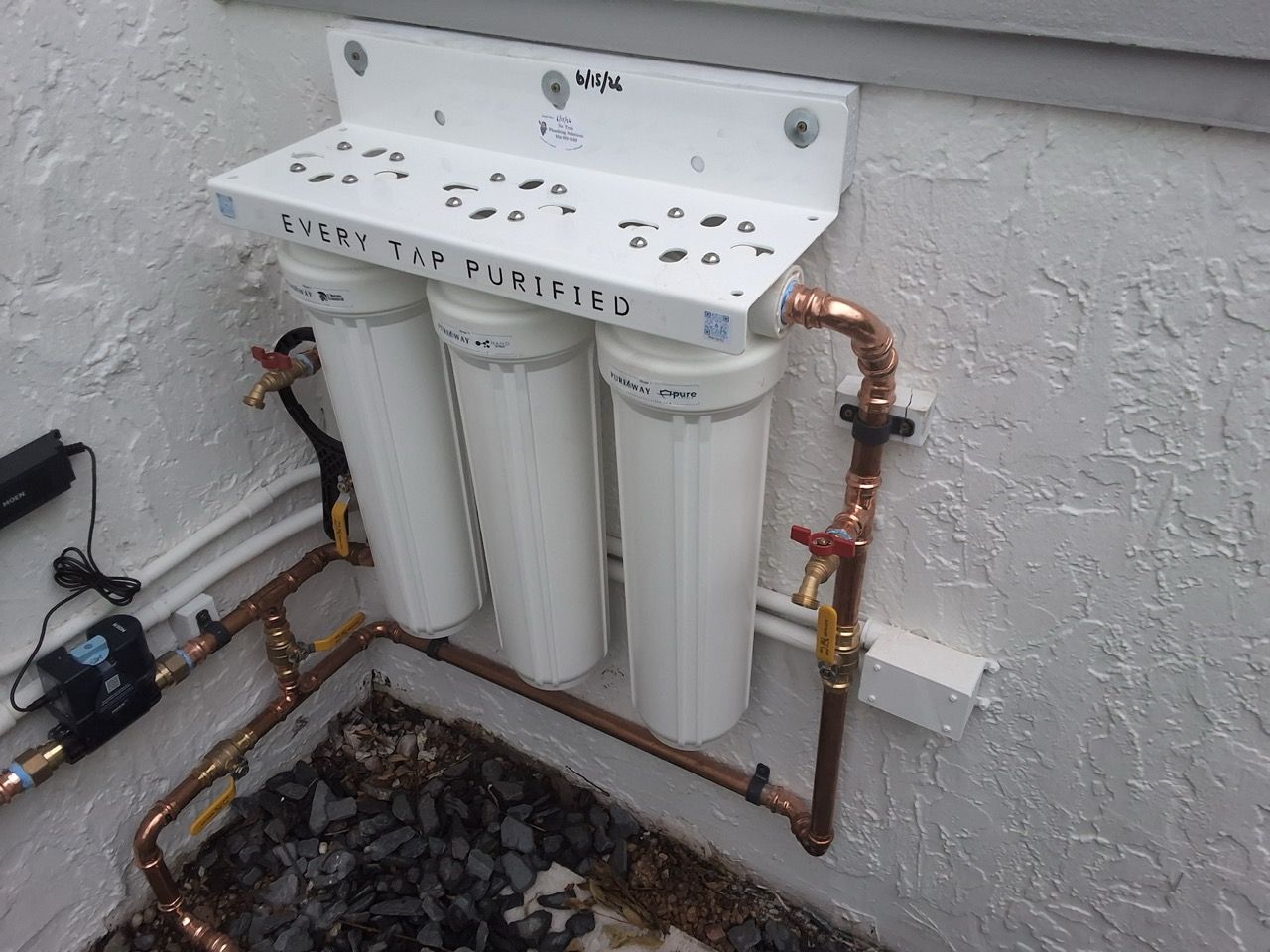 Three white water filter canisters mounted on a wall with copper pipes above a gravel pit