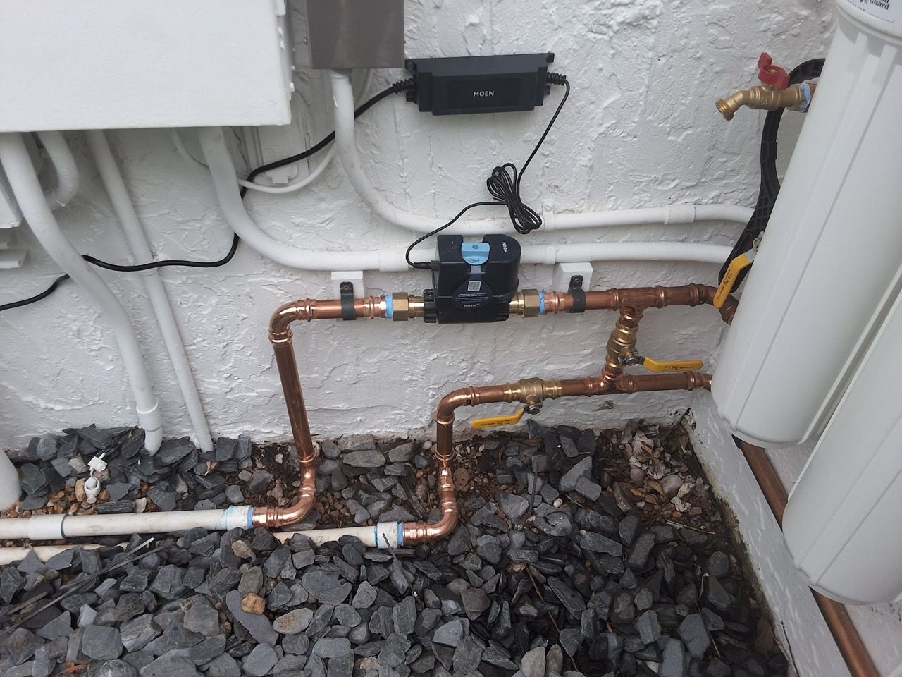 Copper water pipes with a black valve outdoors beside a wall and gravel ground.