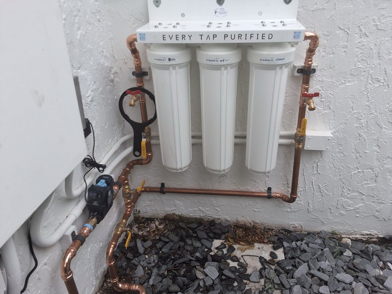 Outdoor wall-mounted water filter system with three white canisters and copper pipes over gravel.