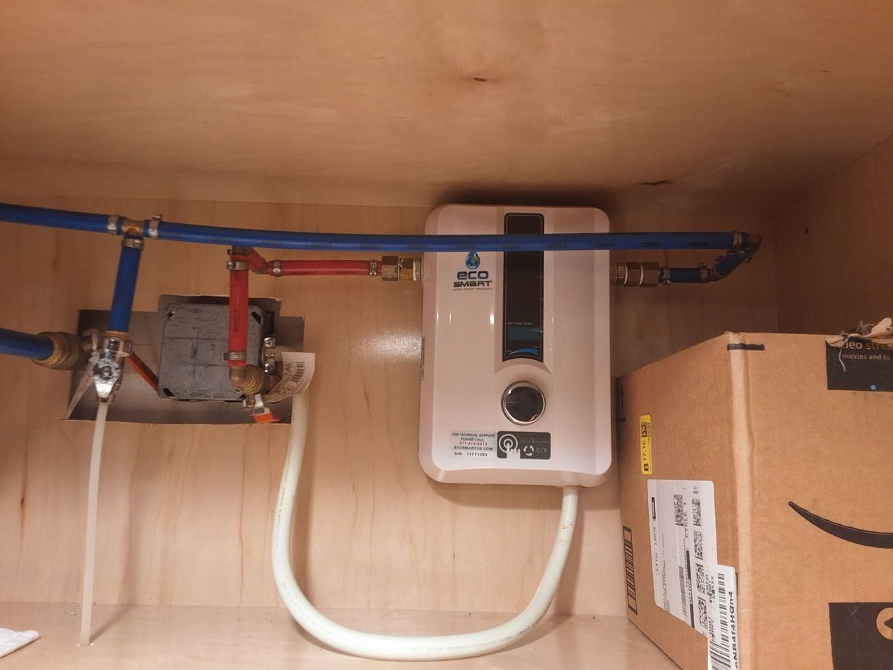 Under-sink water heater with blue and red pipes and a white supply hose inside a cabinet