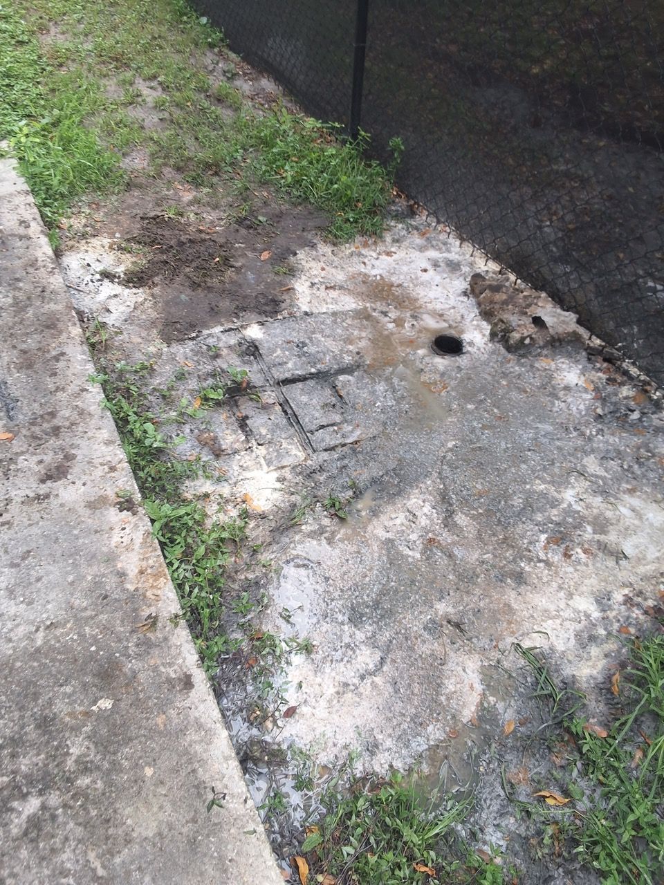 Damaged concrete slab with a hole, surrounded by grass and dirt near a curb