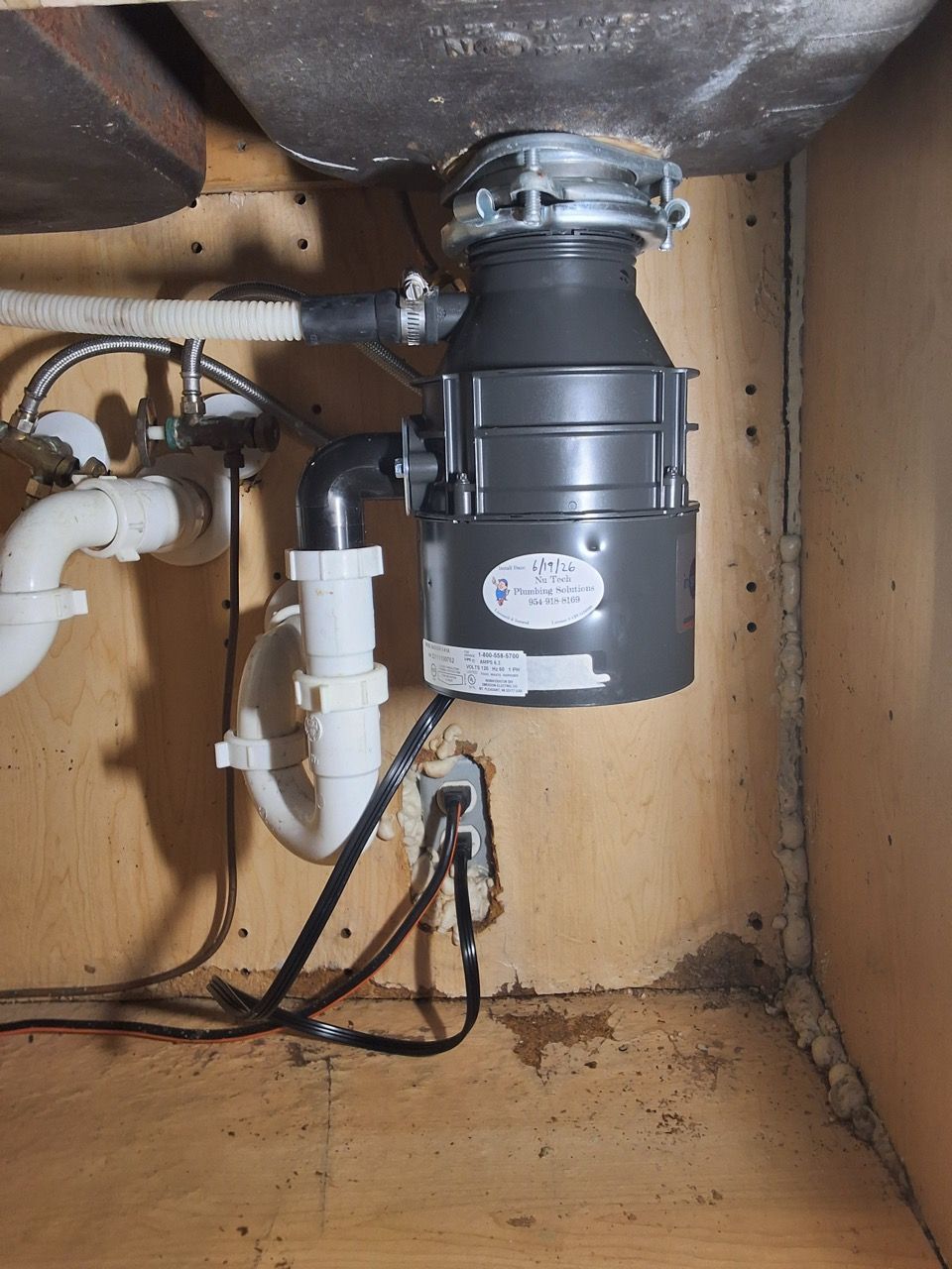 Under-sink garbage disposal and plumbing inside a cabinet, with black and white pipes and a damp wooden base.