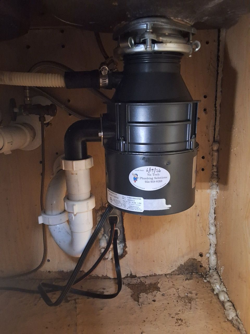 Under-sink garbage disposal unit with plumbing pipes and black drain hoses in a cabinet.