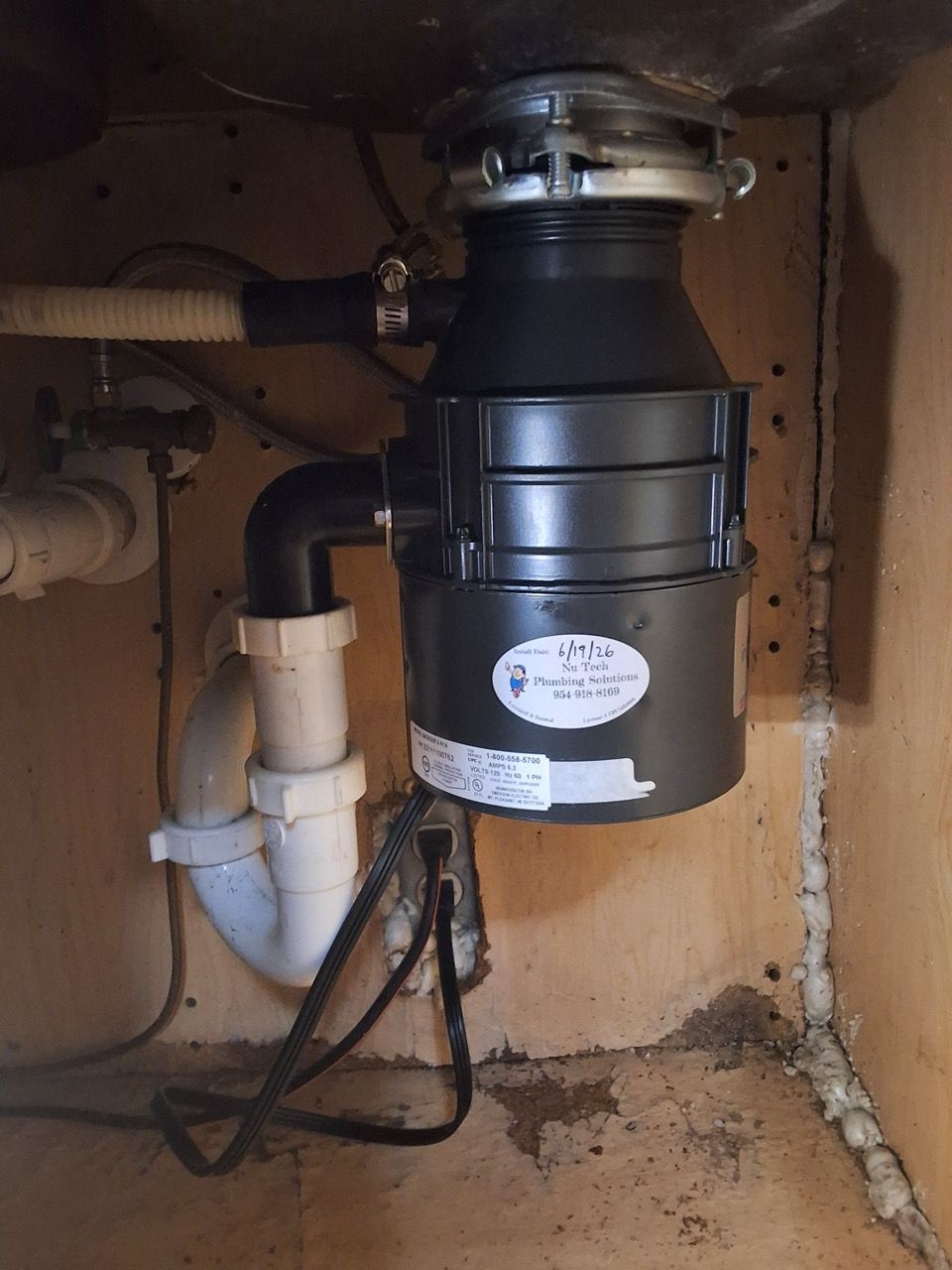 Black garbage disposal under a sink with exposed pipes and wires in a cabinet.
