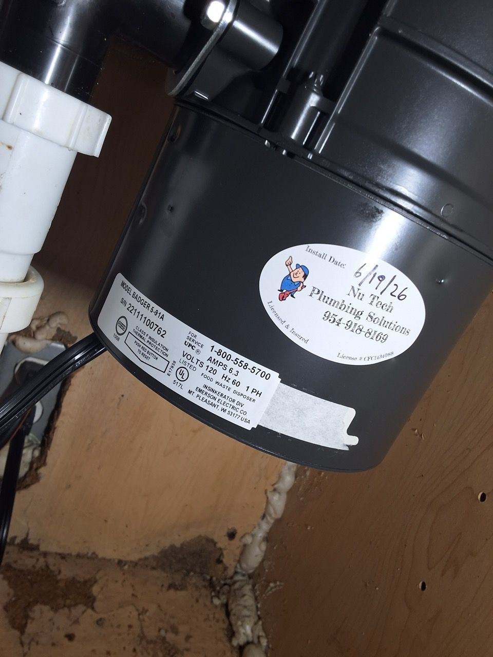 Black garbage disposal under a sink with white pipes and a label, mounted against a wood cabinet interior