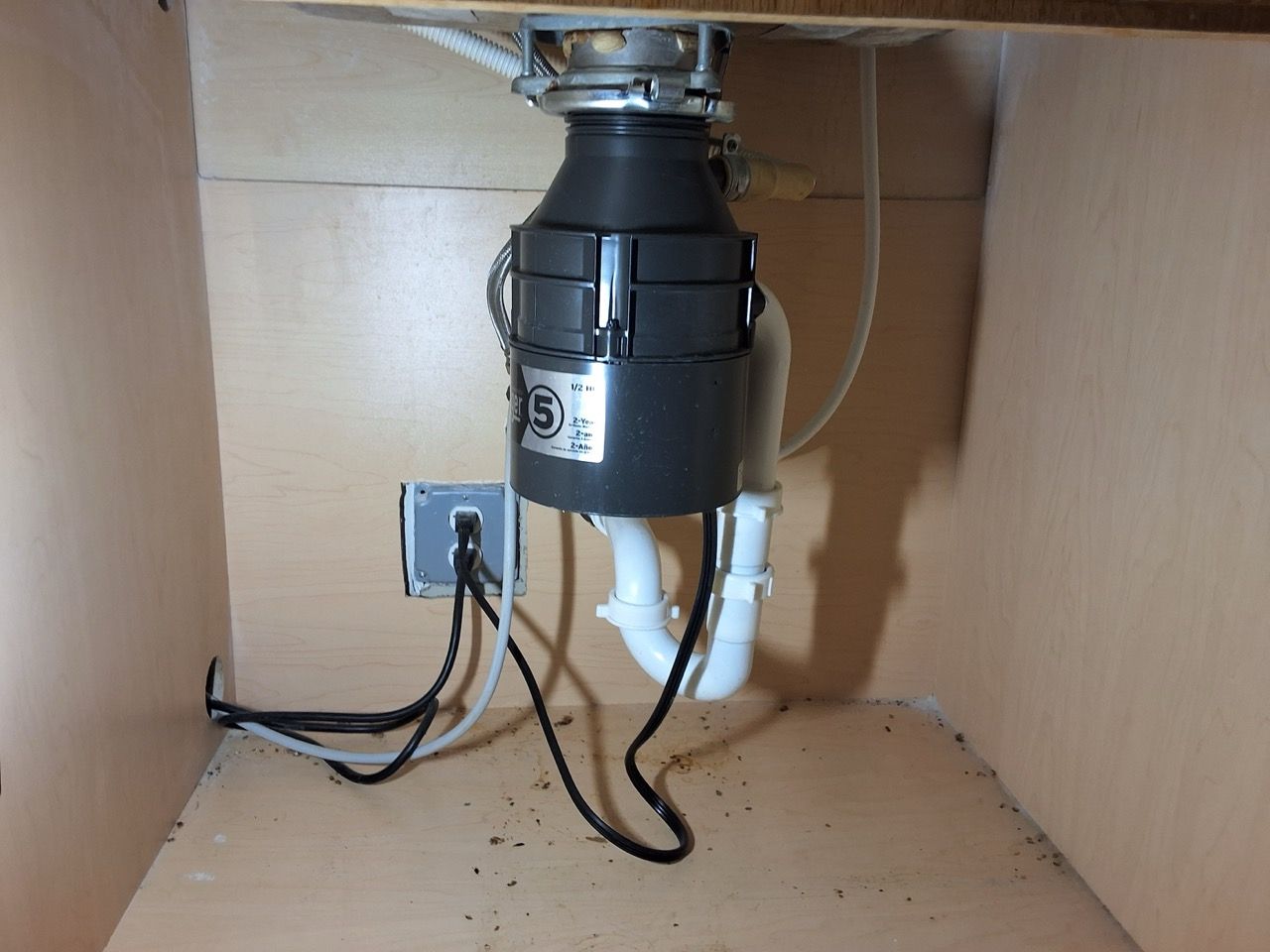 Garbage disposal under a kitchen sink with exposed pipes and a power outlet.