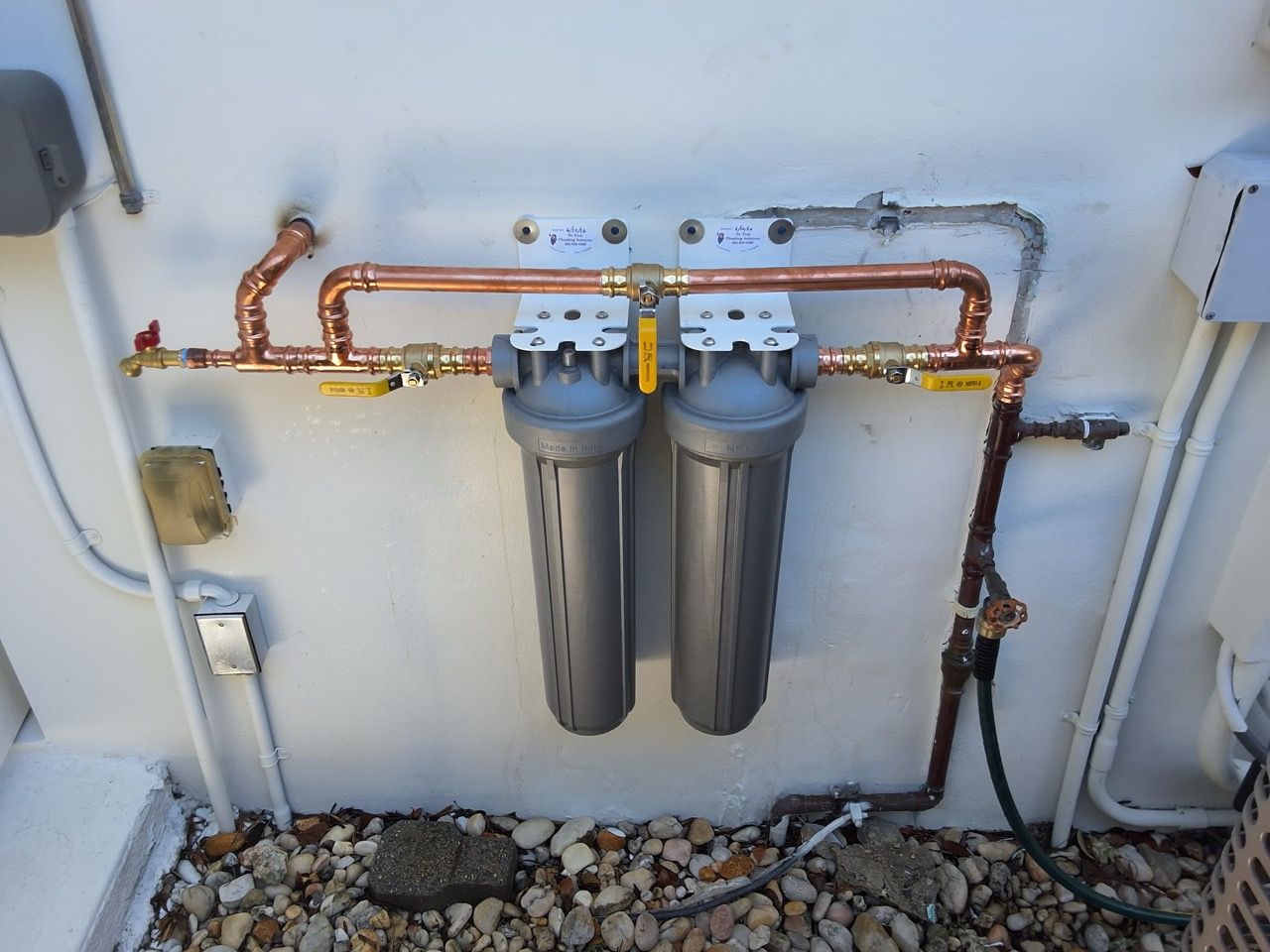 Two gray water filters mounted on a wall with copper pipes and a gravel base below.
