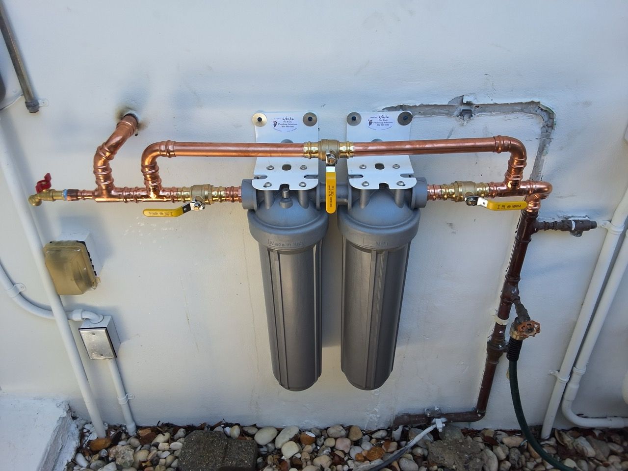 Two gray water filter housings connected to copper pipes on a wall-mounted plumbing system.