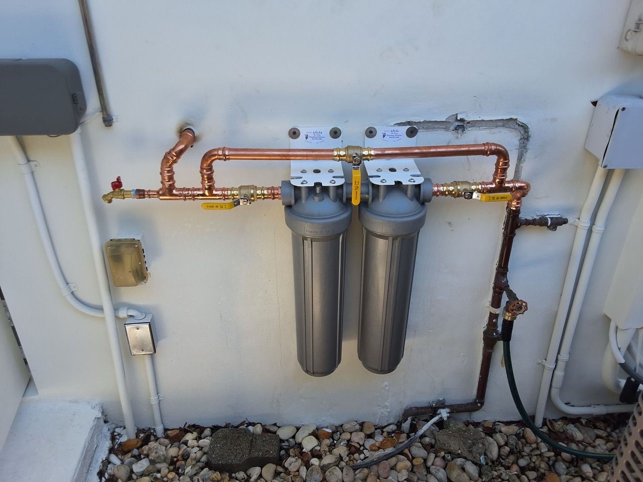 Two water filters with copper piping mounted on an exterior wall above gravel.