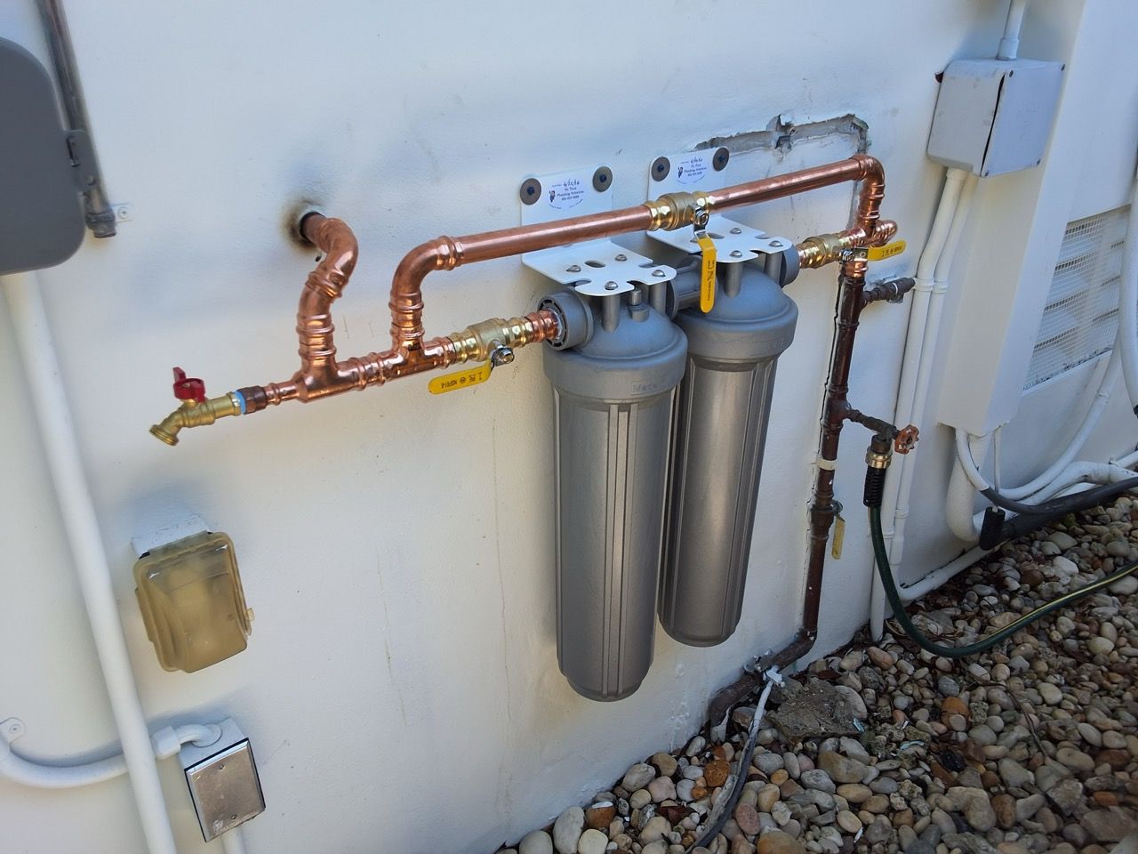 Outdoor water filtration system with copper pipes and two gray filter canisters mounted on a white wall.
