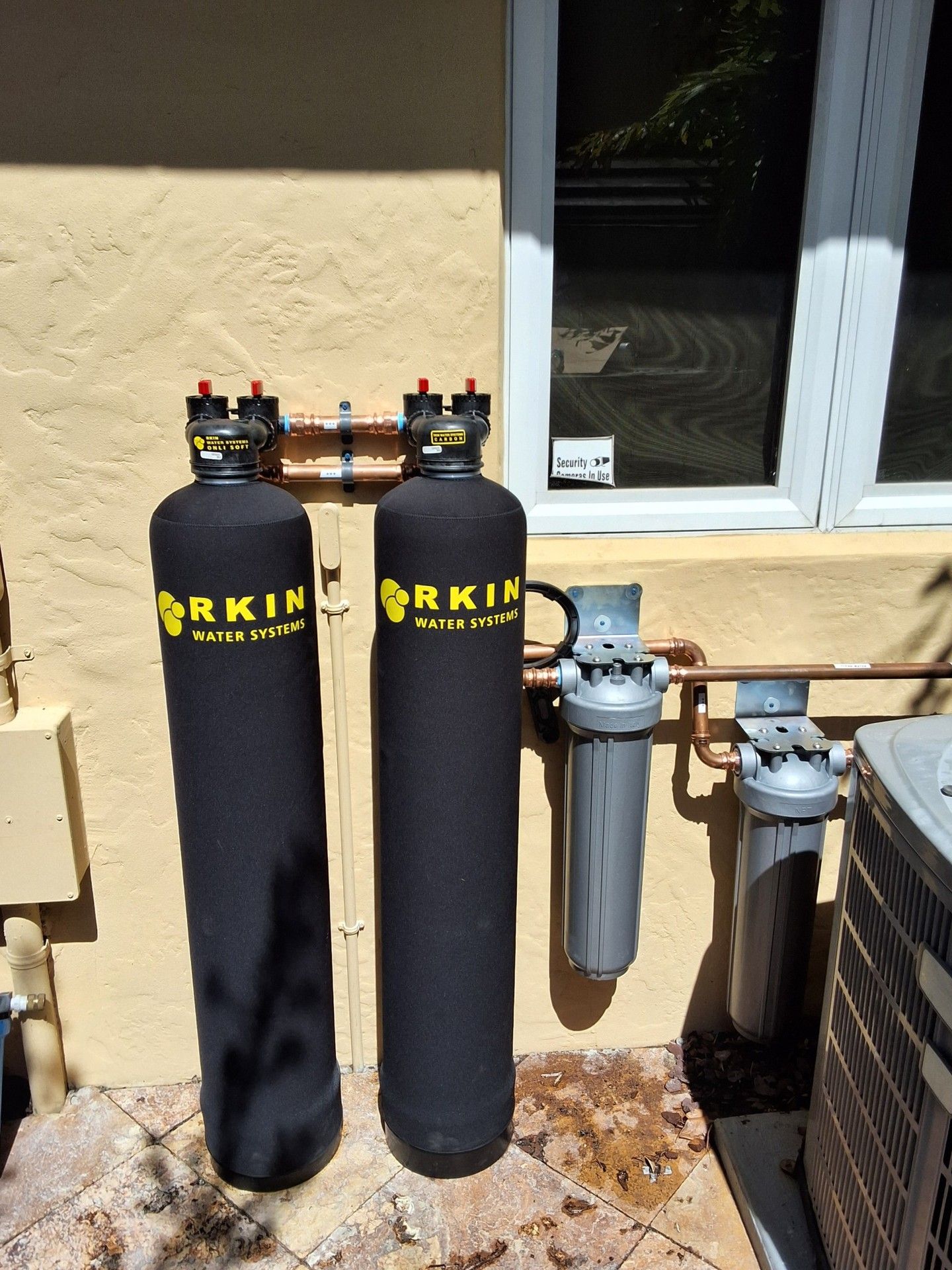 Two black cylindrical filter tanks and two gray water pipes beside a house wall and window