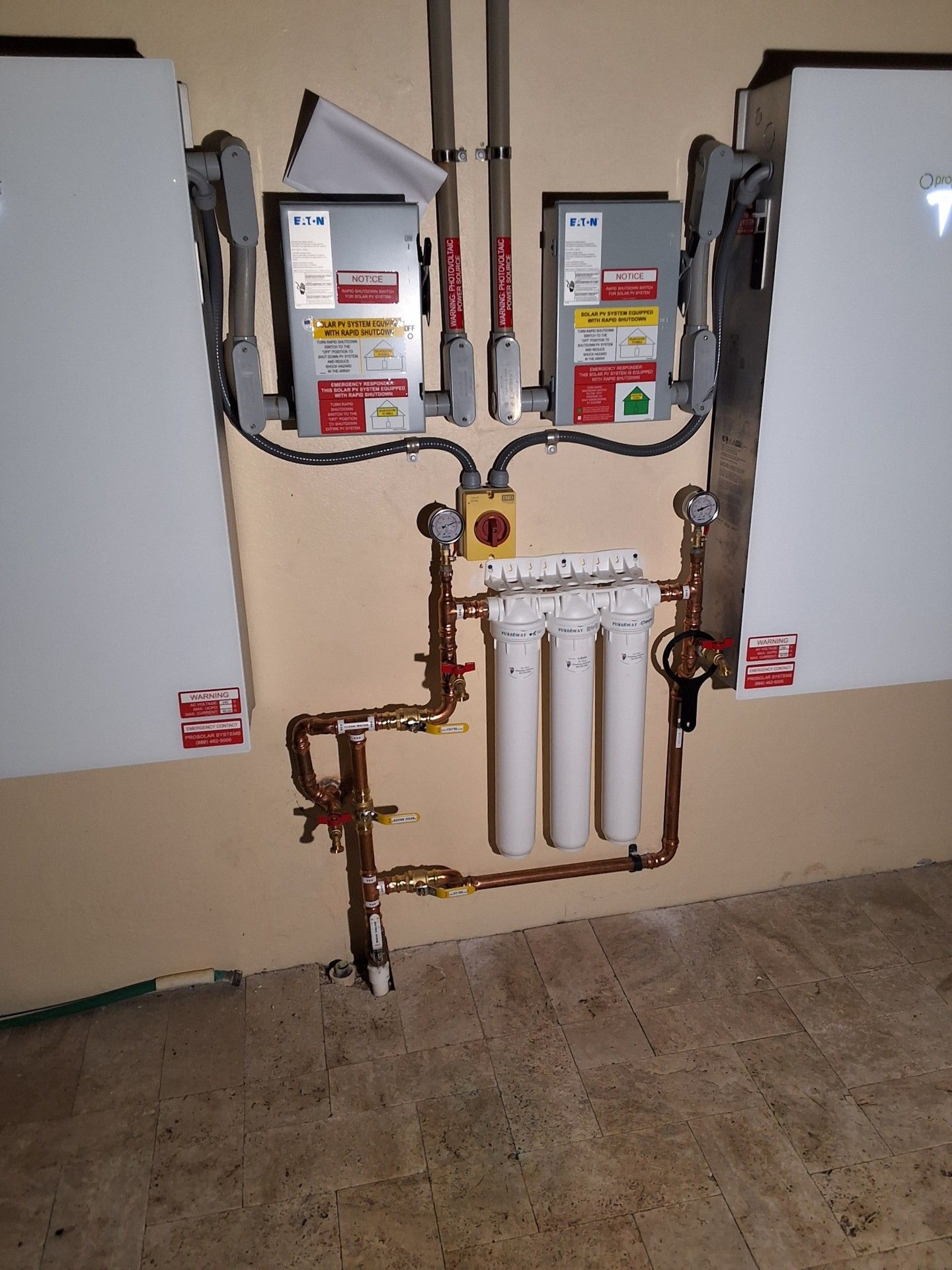 Electrical meter panel with exposed copper pipes and three white filter canisters on a wall indoors